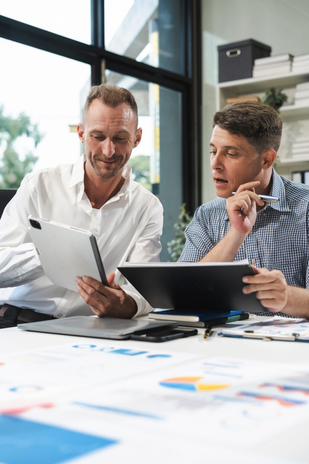 Two businessmen collaborating at a modern office table, reviewing data on a tablet and a laptop with charts spread out in front of them.