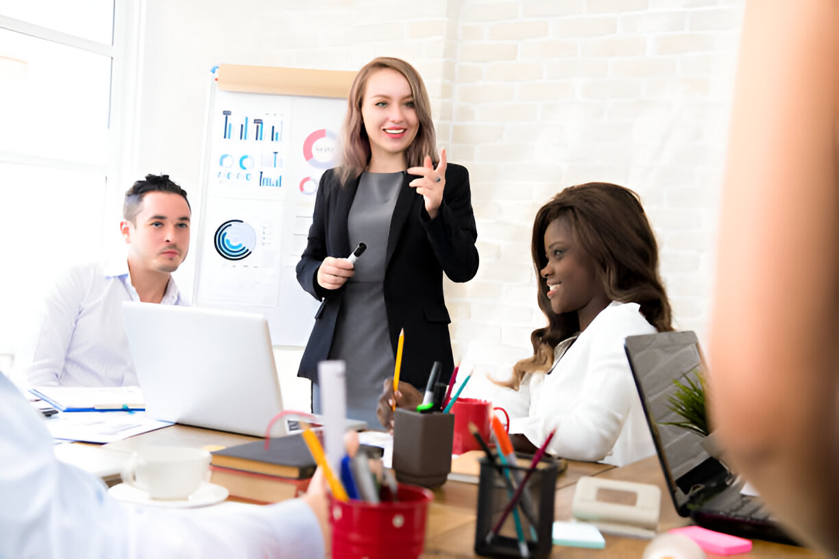 A woman in a black blazer leading a business presentation, speaking to colleagues in a modern office setting with charts and graphs displayed in the background.