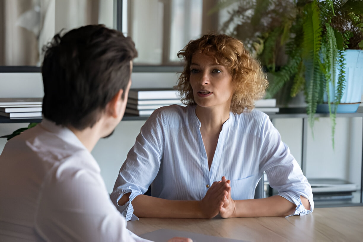 A woman in a light blue blouse having a serious conversation with a man at a table in an office with bookshelves and plants in the background.