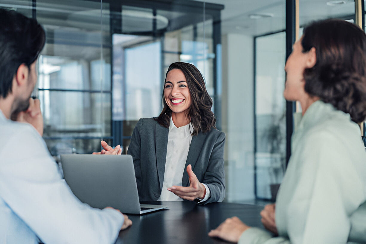 A smiling woman in a business suit gesturing as she talks with two colleagues at a table in a bright office space.