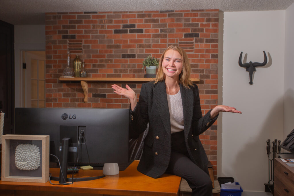 A cheerful woman in a black blazer standing in front of a desk, gesturing with open hands in an office setting with a brick wall background.