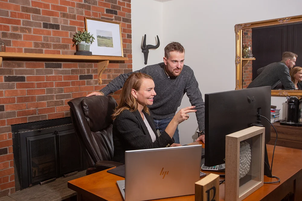 A smiling woman in a black blazer sitting at a desk, pointing at a computer screen while a man stands behind her looking at the screen.
